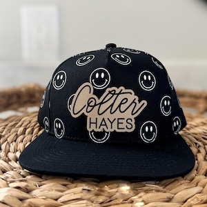 Könnte beinhalten: Schwarze und weiße Baseballkappe mit Smiley-Muster und einem personalisierten braunen Patch mit der Aufschrift "Colter Hayes".
