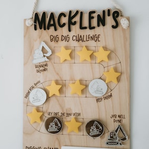May include: A wooden board with a construction theme for a child's reward chart. The board has a title "Macklen's Big Dig Challenge" and features a path with stars and excavator images. The board has a blank space for a reward.