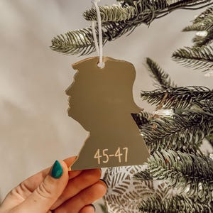 May include: Gold-colored silhouette ornament of a person's head with the number "45-47" written on it. The ornament is hanging on a Christmas tree.