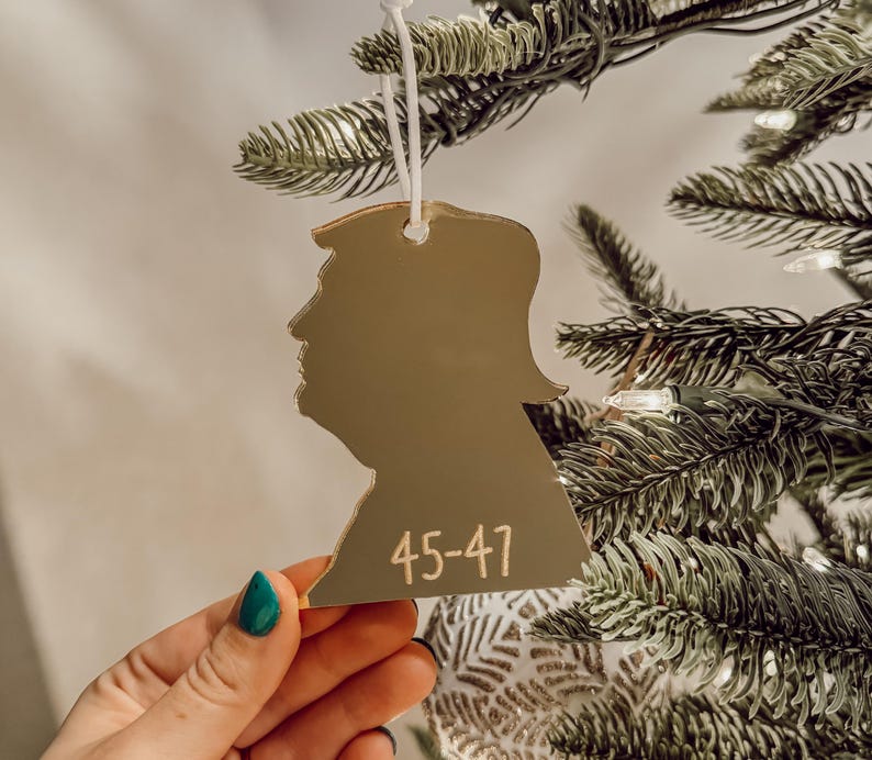 May include: Gold-colored silhouette ornament of a person's head with the number "45-47" written on it. The ornament is hanging on a Christmas tree.