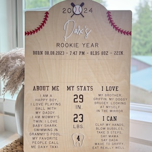 May include: A wooden sign with a baseball theme, celebrating a baby's first year. The sign includes the baby's name, Dax, and details about their birth date, weight, and length. It also includes information about the baby's milestones, such as their height, weight, and number of teeth.