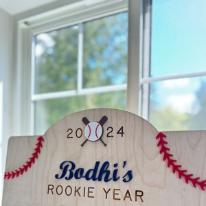 May include: A wooden sign with red stitching around the edges. The sign says "Bodhi's Rookie Year" with a baseball and crossed bats above the text. The year 2024 is written above the baseball.