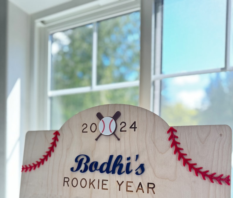 May include: A wooden sign with red stitching around the edges. The sign says "Bodhi's Rookie Year" with a baseball and crossed bats above the text. The year 2024 is written above the baseball.
