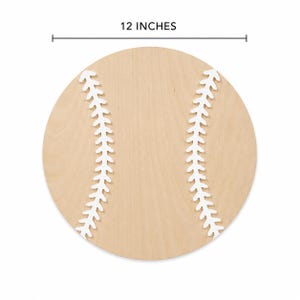 May include: A circular wooden cutout shaped like a baseball, featuring a light tan wood grain finish and two curved white painted lines representing the stitching. The object is labeled with a measurement line indicating a diameter of 12 inches.