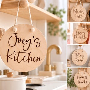 K&ouml;nnte beinhalten: Ein rundes, helles Holzschild mit einer wei&szlig;en Kordel zum Aufh&auml;ngen und dunkelbrauner Schrift. Das Schild zeigt verschiedene personalisierte Namen und Beschriftungen wie Joey's Kitchen, Mack's Hats, Elise's Dress Up, Play Room und Liam's Trucks.