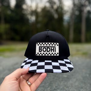 May include: Black and white checkered baseball cap with a black patch that says "BODHI" in white letters.