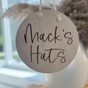 K&ouml;nnte beinhalten: Ein kreisf&ouml;rmiges Schild aus hellem Holz mit der dunkel eingravierten Aufschrift Mack's Hats. Das Schild ist an einem wei&szlig;en Seil aufgeh&auml;ngt, das durch zwei L&ouml;cher an der Oberseite gef&uuml;hrt ist, wobei die Seilenden auf der Vorderseite verknotet sind.