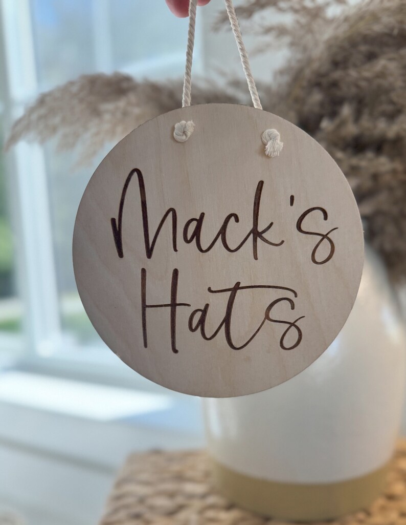 K&ouml;nnte beinhalten: Ein kreisf&ouml;rmiges Schild aus hellem Holz mit der dunkel eingravierten Aufschrift Mack's Hats. Das Schild ist an einem wei&szlig;en Seil aufgeh&auml;ngt, das durch zwei L&ouml;cher an der Oberseite gef&uuml;hrt ist, wobei die Seilenden auf der Vorderseite verknotet sind.