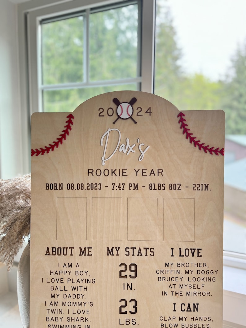 May include: A wooden sign with a baseball theme, celebrating a baby's first year. The sign features the baby's name, "Dax's", and the date of birth, "08.08.2023". It also includes the baby's stats, such as height, weight, and other milestones.