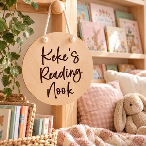 K&ouml;nnte beinhalten: Ein rundes, helles Holzschild, das an einem wei&szlig;en Seil h&auml;ngt. Das Schild zeigt den dunklen, eingravierten Text "Keke's Reading Nook" in Schreibschrift. Es ist mit zwei kleinen wei&szlig;en Kn&ouml;pfen oben an der runden Holzplatte befestigt.
