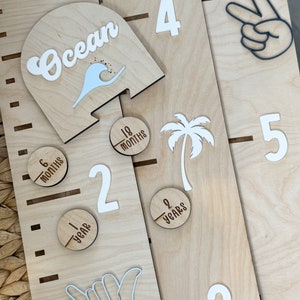 May include: A wooden growth chart with a beach theme. The chart features a surfboard with the word "Ocean" on it, a palm tree, and hand shapes. The chart is marked with numbers and months, including 6 months, 18 months, 1 year, 2 years, 3, 4, and 5.