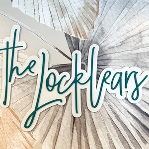 May include: A teal green cursive script sign with the text "the locklear's" on a white background with a watercolor design of brown and gray palm leaves.