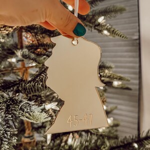 May include: A gold-colored, cutout ornament shaped like a person's head with the number "45-47" inscribed on the bottom. The ornament is hanging from a white string on a Christmas tree.