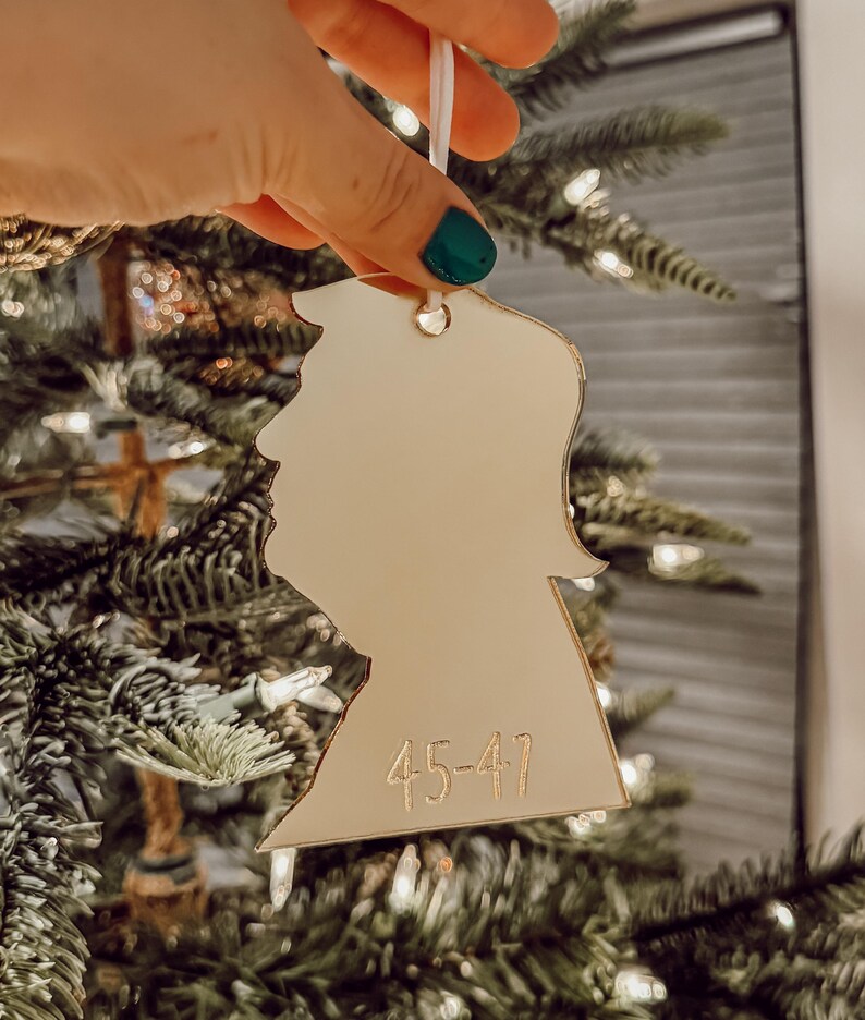 May include: A gold-colored, cutout ornament shaped like a person's head with the number "45-47" inscribed on the bottom. The ornament is hanging from a white string on a Christmas tree.
