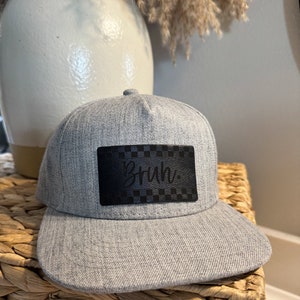 May include: Gray and black baseball cap with a black leather patch that says "Bruh" in white cursive lettering.