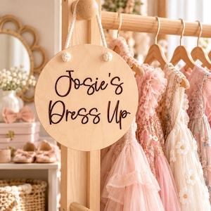 K&ouml;nnte beinhalten: Ein rundes, helles Holzschild, das an einer Kleiderstange aus Holz h&auml;ngt, mit dem Text "Josie's Dress Up" in dunkelbrauner Schreibschrift. Das Schild ist an einem wei&szlig;en Seil befestigt und h&auml;ngt vor mehreren Kleidern auf Kleiderb&uuml;geln.