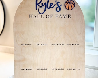 May include: A wooden sign with an arched top, painted in a light wood color. The sign has the text "Kyle's Hall of Fame" written in black lettering. There is a basketball graphic in orange and black on the top right corner of the sign. The sign is divided into 12 sections, each labeled with a month of the year.