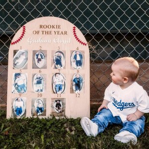 以下が含まれることがあります： 野球をテーマにした木製看板で、「Rookie of the Year Graham Cloyd」の文字が書かれています。看板には12個の番号が付けられた正方形があり、それぞれに赤ちゃんの写真が貼られています。白い野球のユニフォームを着て、「Dodgers」と書かれた帽子をかぶった赤ちゃんが看板の前に座っています。