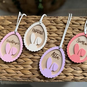 Könnte beinhalten: Vier eiförmige Holzanhänger mit Namen und Hasenohren. Die Anhänger sind in Rosa, Weiß und Lila gehalten und tragen die Namen "Hudsyn", "Farrah", "Atta" und "Sierra" in Schreibschrift. Jeder Anhänger hat eine weiße Schnur.