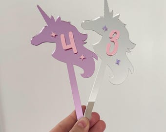 May include: Two unicorn shaped cake toppers, one silver and one pink with a pink number 3 and a pink number 4 on them.