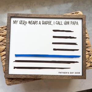 May include: A rectangular wooden sign with a white background featuring the black text MY HERO WEARS A BADGE, I CALL HIM PAPA and FATHER'S DAY 2025. The design includes horizontal black brush strokes and a single horizontal blue line across the center.