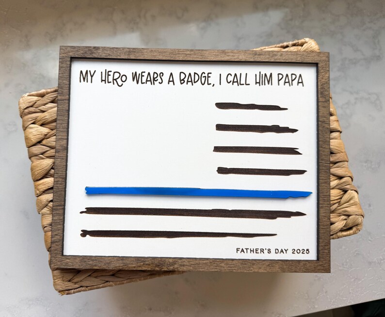 May include: A rectangular wooden sign with a white background featuring the black text MY HERO WEARS A BADGE, I CALL HIM PAPA and FATHER'S DAY 2025. The design includes horizontal black brush strokes and a single horizontal blue line across the center.