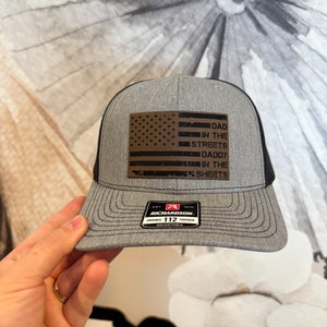 May include: Gray and black trucker hat with a brown leather patch featuring an American flag and the text "Dad in the Streets Daddy in the Sheets". The hat has a red, white, and black Richardson logo with the text "Original 112 Trucker Adjustable".