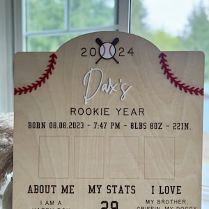 May include: A wooden sign with a baseball theme, announcing a baby's "Rookie Year." The sign includes the baby's name, "Dax's," birthdate, time, weight, and length. The sign also includes the text "About Me," "My Stats," and "I Love." The sign is decorated with red stitching and a baseball and bats.