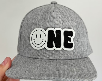 May include: Gray baseball cap with a black and white smiley face and the word "ONE" in black letters.