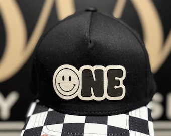 May include: A black baseball cap with a flat brim featuring a black and white chequerboard pattern. The front of the cap displays a white patch with the black text "ONE" and a smiley face replacing the letter "O". The text reads "Shown: White patch + Black text".