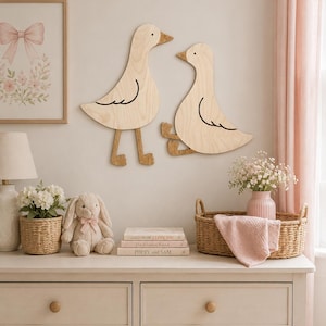 May include: Two light-colored wooden wall decorations shaped like ducks, one standing and one sitting, featuring simple black line details for wings and eyes. The ducks are mounted on a wall above a dresser with various decorative items.