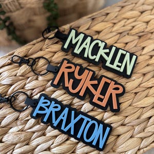 May include: Three personalized keychains with names: MACKLEN in green, RYKER in orange, and BRAXTON in blue. Each keychain has a black background and a black clasp. The keychains are displayed on a woven wicker surface.