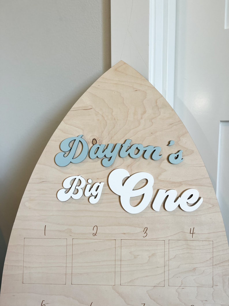May include: A wooden surfboard-shaped sign with the text "Dayton's Big One" in blue and white lettering. The sign has four blank squares numbered 1 through 4.