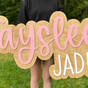 May include: A wooden sign with the name "Tayslee Jade" written in pink and white paint. The sign is held by a person in front of a green grassy background.