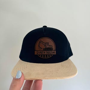 May include: Black and tan baseball cap with a brown leather patch featuring a cartoon excavator and the text "Bowen Mason".