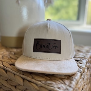 May include: A beige and brown baseball cap with a brown leather patch that says "Braxton" in black cursive lettering.