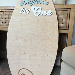 May include: A wooden surfboard-shaped sign with the text "Dayton's Big One" in blue and white lettering. The sign has 12 numbered squares for photos or other decorations. A wave design is at the bottom of the sign.