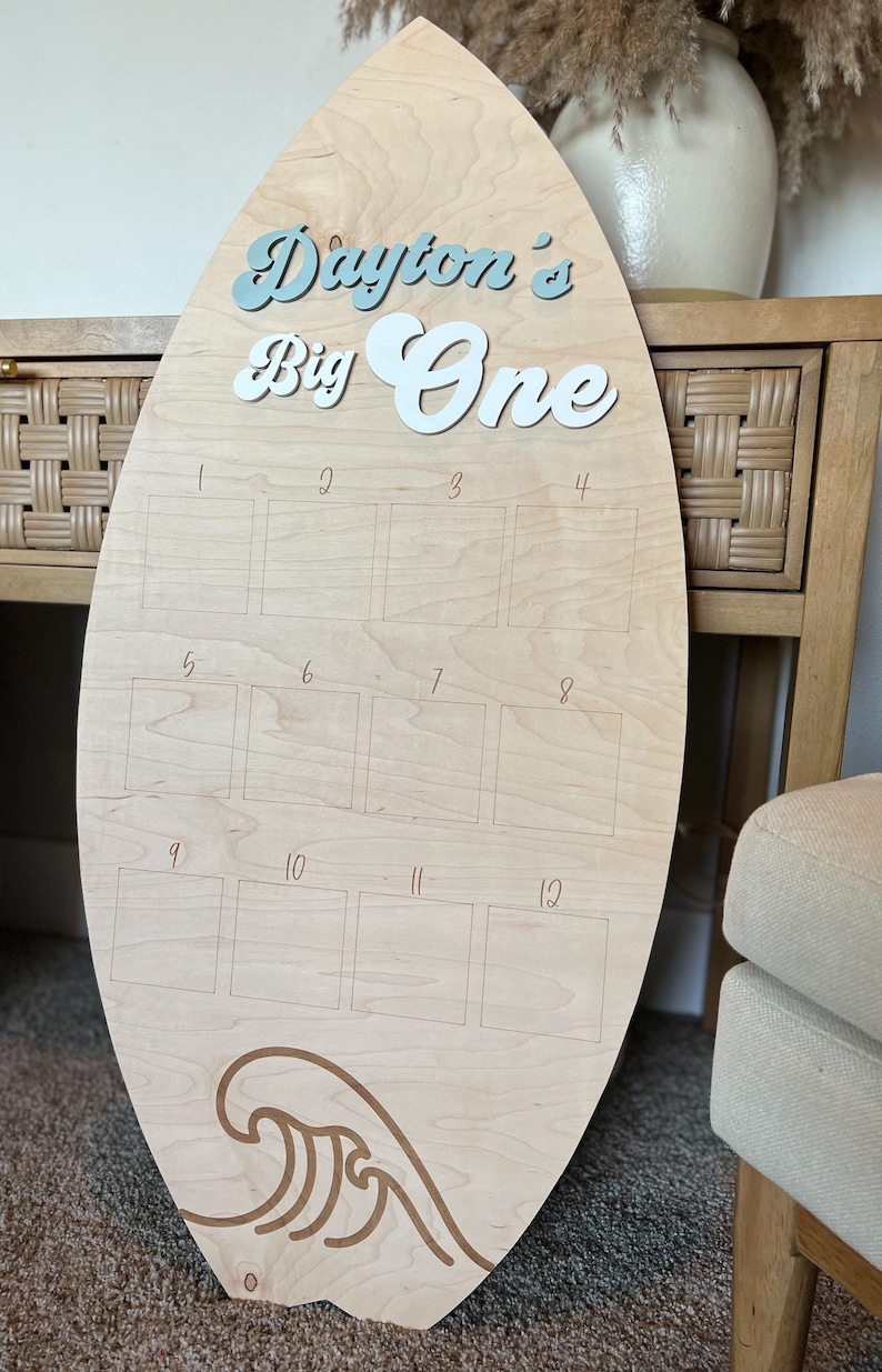 May include: A wooden surfboard-shaped sign with the text "Dayton's Big One" in blue and white lettering. The sign has 12 numbered squares for photos or other decorations. A wave design is at the bottom of the sign.