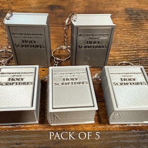 May include: A collection of five silver-colored book-shaped keychains. Each keychain features the words "Holy Scriptures" on the cover. The text "Pack of 5" is visible at the bottom of the image.
