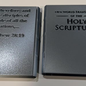 May include: Two gray, book-shaped objects. The left one has the text "Go, therefore; and make disciples of people of all the nations,... Matthew 28:19". The right one reads "NEW WORLD TRANSLATION OF THE HOLY SCRIPTURES".