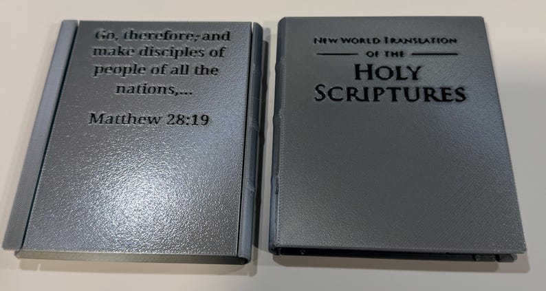 May include: Two gray, book-shaped objects. The left one has the text "Go, therefore; and make disciples of people of all the nations,... Matthew 28:19". The right one reads "NEW WORLD TRANSLATION OF THE HOLY SCRIPTURES".