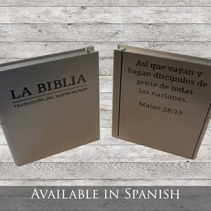 May include: Two beige-colored books are displayed, one open and one closed. The closed book has the text "LA BIBLIA" and "TRADUCCI&Oacute;N DEL NUEVO MUNDO" printed on the cover. The open book displays a quote from Matthew 28:19 in Spanish.