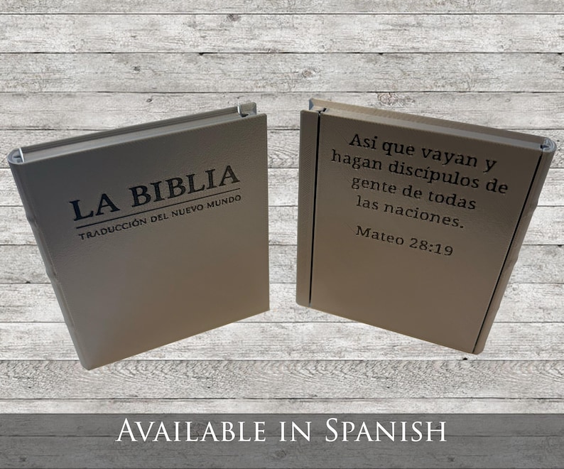May include: Two beige-colored books are displayed, one open and one closed. The closed book has the text "LA BIBLIA" and "TRADUCCI&Oacute;N DEL NUEVO MUNDO" printed on the cover. The open book displays a quote from Matthew 28:19 in Spanish.