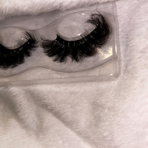 May include: A pair of black, voluminous false eyelashes presented in a clear plastic case. The lashes are thick and appear fluffy. The case is set against a white, textured background, providing a contrast between the clear plastic and the soft surface.