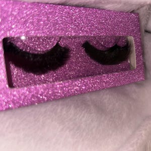 Può includere: Un paio di ciglia finte nere esposte in una scatola color magenta scintillante. La scatola ha una finestra rettangolare che mostra le ciglia. Lo sfondo è un tessuto morbido, bianco e strutturato.