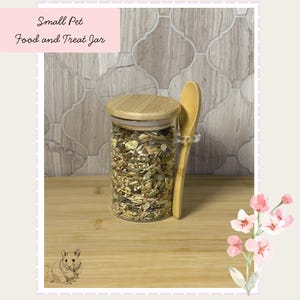 Op de afbeelding: Een doorzichtige glazen pot gevuld met een mix van zaden en granen, voorzien van een rond bamboe deksel en een houten lepel aan de zijkant. De tekst Small Pet Food and Treat Jar is zichtbaar. De pot staat op een houten ondergrond.