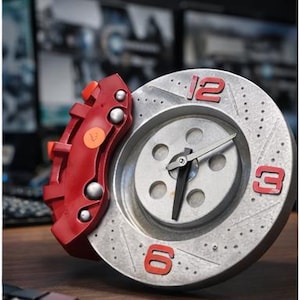 May include: A silver and red automotive-themed clock. The clock face is designed to resemble a brake rotor, with red numerals at 12, 3, and 6 o'clock. A red brake caliper is attached to the side. The clock is on a wooden surface.