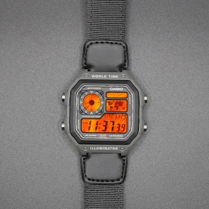 May include: A digital Casio watch with a gray case and a black nylon strap. The watch face displays the time in orange digits, along with other functions. The watch has buttons for mode, adjust, search, and light. The watch is on a gray surface.