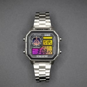 May include: A silver Casio digital watch with a rectangular face and a matching stainless steel band. The watch face displays the time, date, and world time features. The watch has the text "CASIO", "WORLD TIME", and "ILLUMINATOR".
