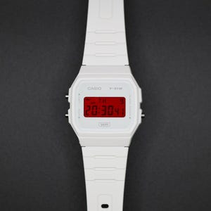 May include: White Casio F-91W digital watch with a rectangular face and a matching white strap. The watch displays the time in red digits. The watch has the Casio logo and model number printed on the face.